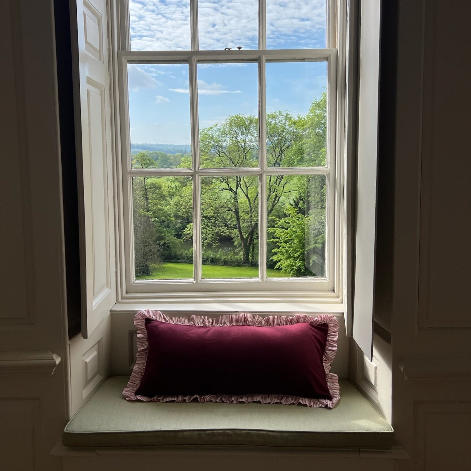 Window with a view of english countryside and a burgandy ruffle pillow on a windowsill.