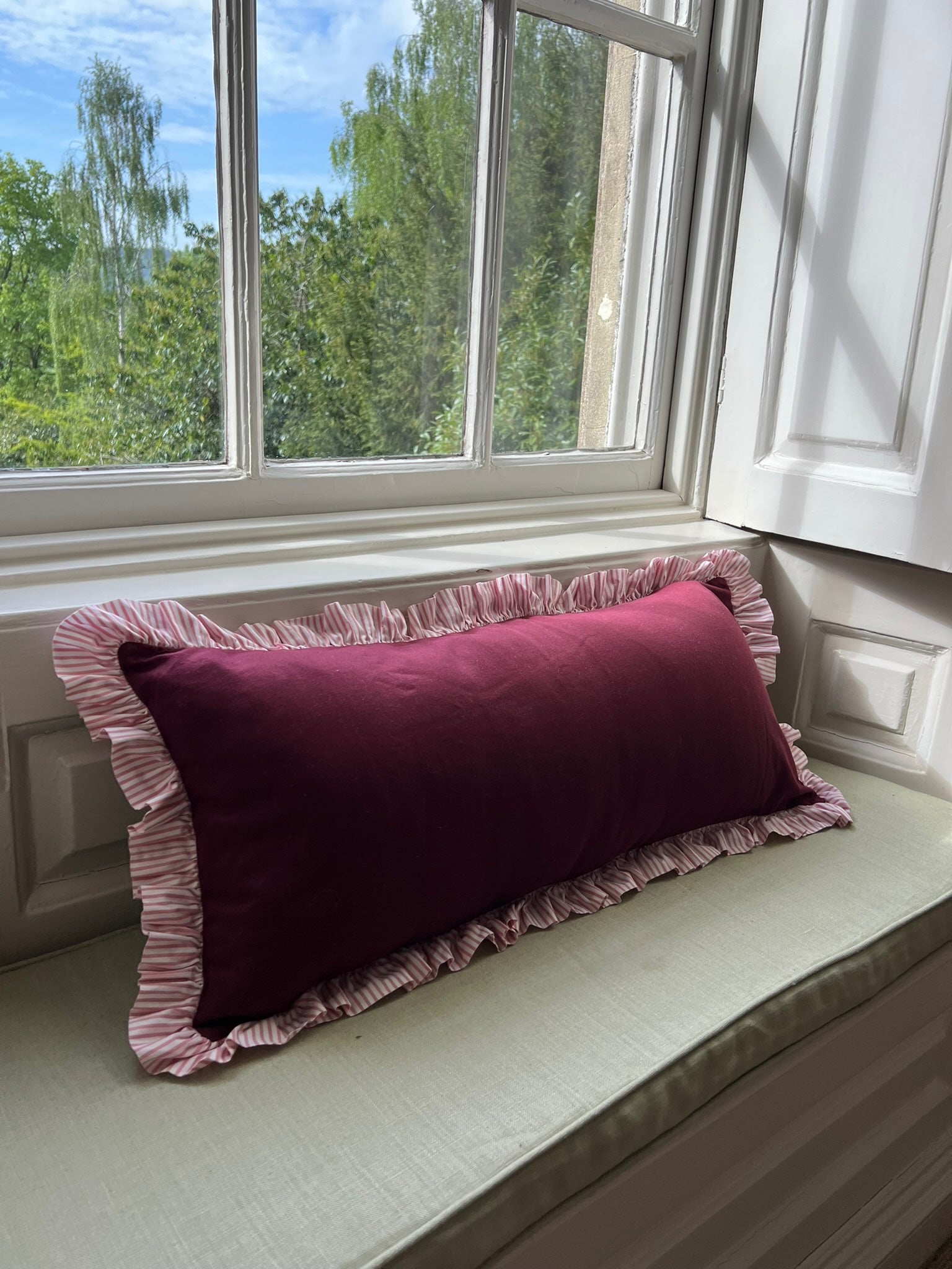 Burgandy pillow with pink ruffle on a windowsill with a view of english countryside